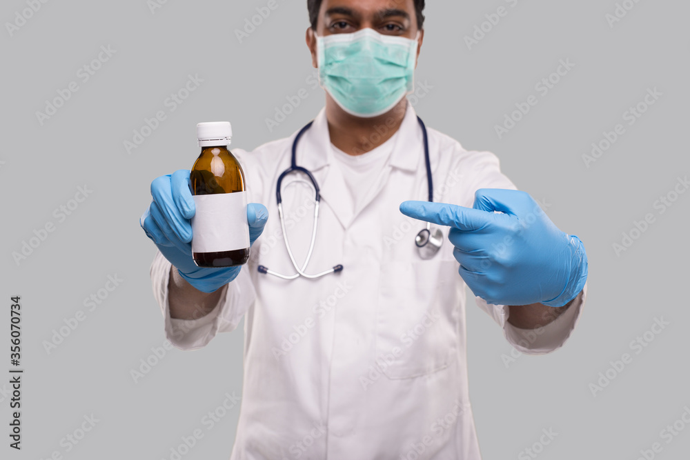 Indian Man Doctor Pointing at Cough Syrup Wearing Medical Mask and Gloves. Hands Wash Antiseptic. Corona Virus Concept. Isolated Indian Man Doctor Pointing at Cough Syrup Wearing Medical Mask and Gloves. Hands Wash Antiseptic. Corona Virus Concept. Isolated