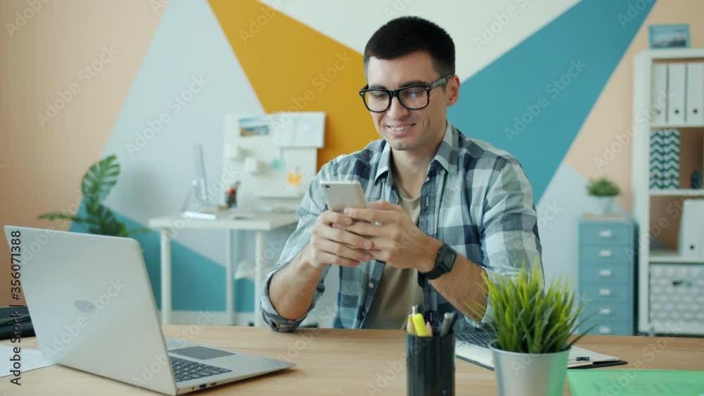 Handsome young man is using smartphone in office at desk enjoying communication online touching screen. Modern technoology and millennials concept. Handsome young man is using smartphone in office at desk enjoying communication online touching screen. Modern technoology and millennials concept.
