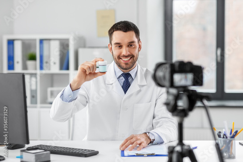 healthcare, medicine and blogging concept - happy smiling male doctor with camera showing drug and recording video blog at hospital