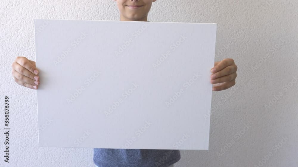 Boy holding in his hands and show blank empty white mock up board poster. Grey wall on background. Close up