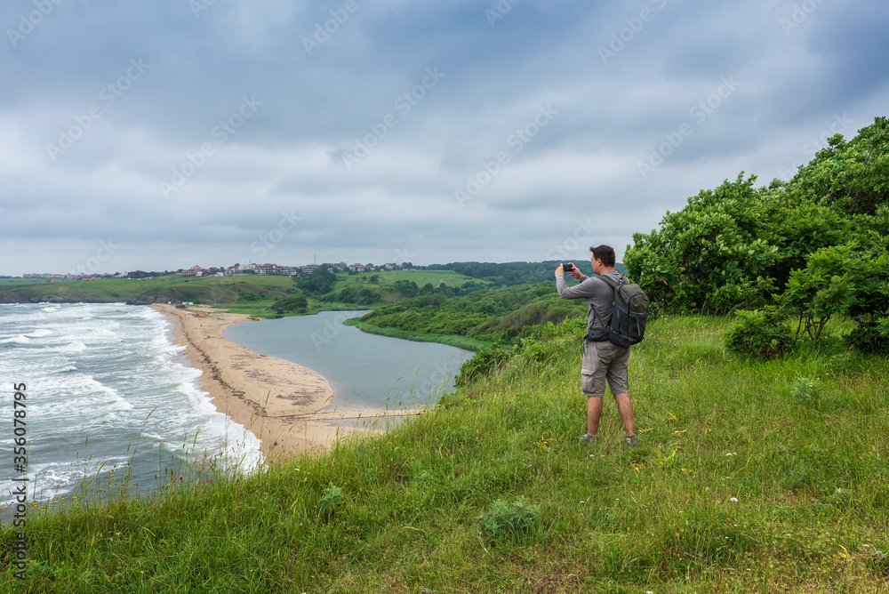 Rear view of a man taking pictures on his smart phone while standing at the edge of a rock looking toward beautiful and wild beach at the Black Sea coast and the estuary of Veleka river, Bulgaria.