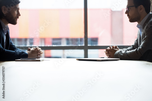 Side view two confident serious businessmen with tensed nerves sit at boardroom desk looking at each other, rivalry bad attitude, confrontation during difficult negotiation, leadership tussle concept