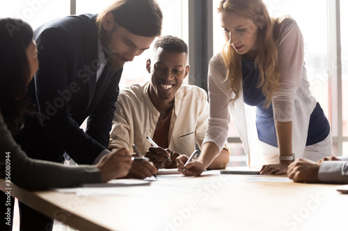 Diverse associates gathered in boardroom leaned over desk writing project plan and ideas do paperwork at group meeting briefing, analyzing market, having fun, teamwork, fun team building games concept