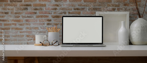 Home office table with mock-up laptop, supplies and decorations on white desk with brick wall