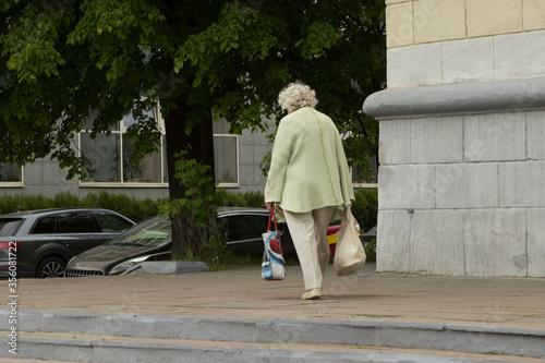 Bild auf Leinwand A lonely old gray-haired woman carries bags and returns home from the store, walks along the city street in the summer