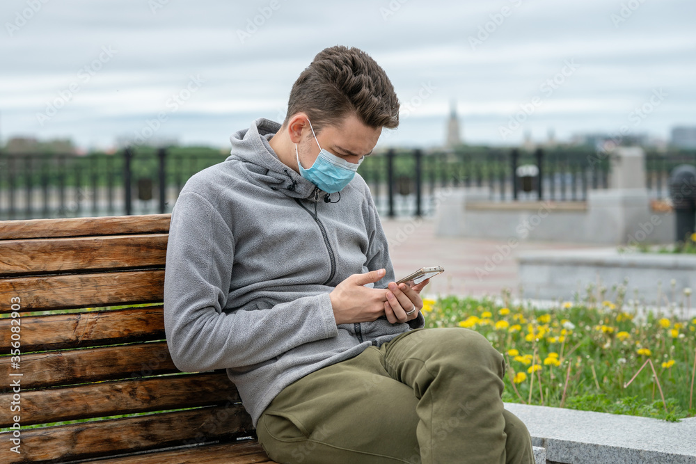 Obraz premium Young man on the street sits on a bench in a medical mask and with a phone in his hands.