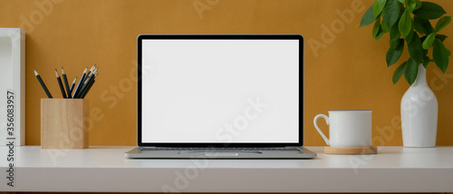 Creative worktable with mock-up laptop, stationery, cup and decorations on white table with yellow wall