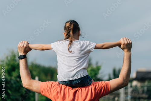 Father holding his daughter on the shouldders and standing on nature summer background. Family or fathers day concept. Back view.