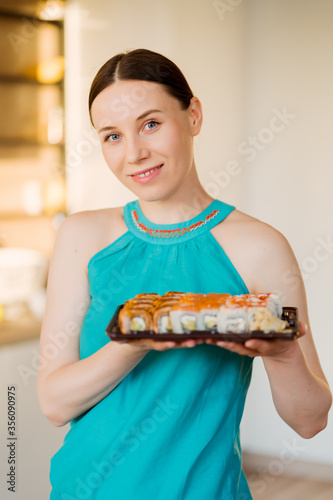 Portrait charismatic woman presenting a plate with sushi rolls in her hands with modern kitchen in the background