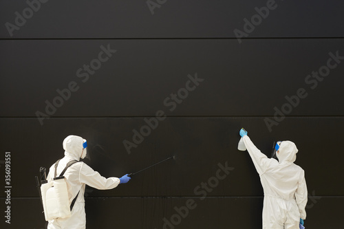 Wide angle view at two workers wearing protective gear and spraying chemicals over black building facade during disinfection or cleaning, copy space photo