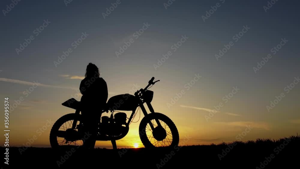 A girl near the bike watches the sunset and enjoys life, in the fresh air in the middle of a field in nature, a biker with a retro motorcycle.