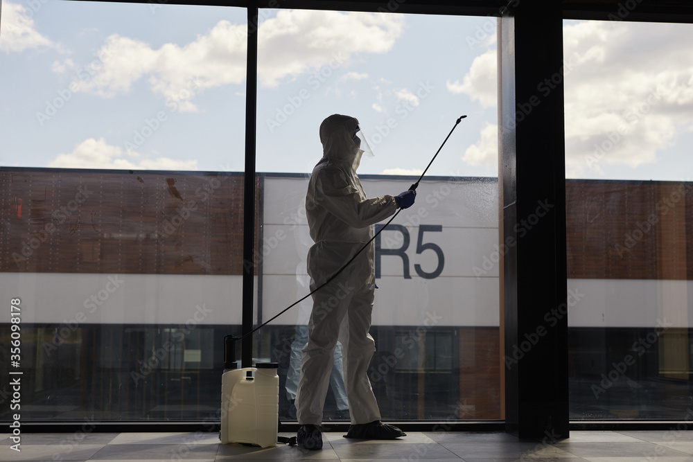 Backlit full length portrait of worker wearing protective suit spraying chemicals during disinfection or cleaning, copy space