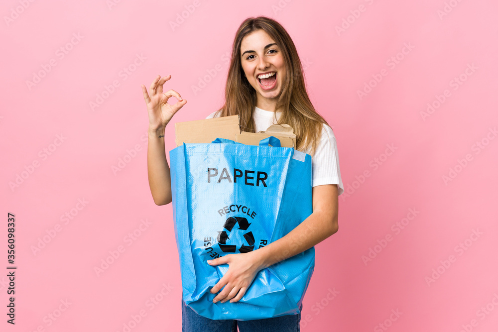 holding a recycling bag full of paper to recycle over isolated pink showing an ok sign with fingers