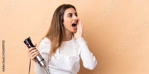 Woman using hand blender over isolated background shouting with mouth wide open to the side