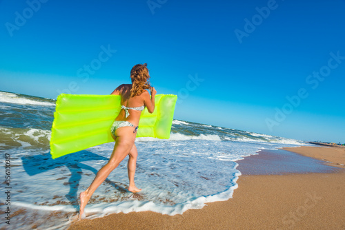 Beautiful young slim woman in a swimsuit runs along the beach with an air mattress along the sandy shore near the stormy sea waves on a sunny warm summer day. Tourism and vacation concept