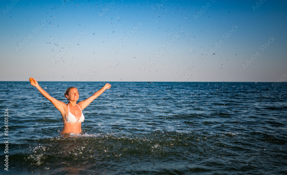 Beautiful cheerful young woman bathes in water and rejoices raising her hands up on sunny warm summer evening. Concept of enjoying a long-awaited vacation. Copyspace