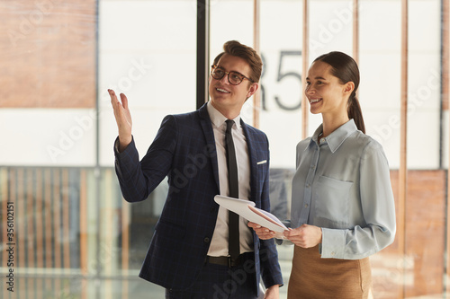 Waist up portrait of smiling real estate agent discussing property with female client and pointing up while standing in empty office building interior lit by sunlight, copy space photo