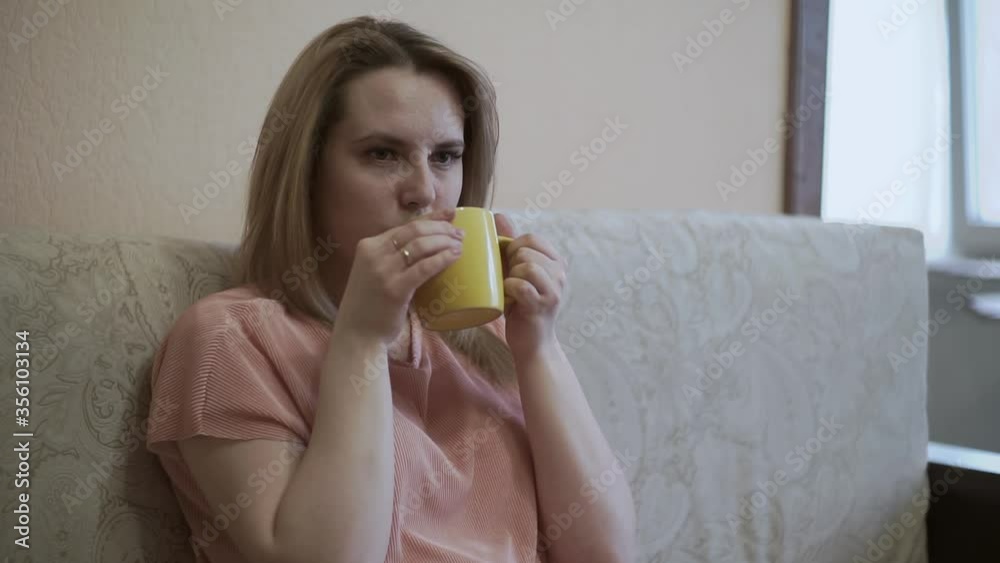 Beautiful girl is sitting at home in quarantine. Picks up a remote control, drinks hot drink tea, watches TV, enjoys spending free time sitting sofa living room in apartment Beautiful girl is sitting at home in quarantine. Picks up a remote control, drinks hot drink tea, watches TV, enjoys spending free time sitting sofa living room in apartment