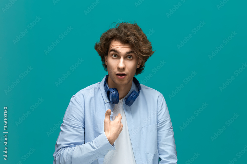 are you blame me. Portrait of a young beautiful man wearing white t-shirt and blue shirt in blue headphones points himself by finger and looking at the camera are you blame me. Portrait of a young beautiful man wearing white t-shirt and blue shirt in blue headphones points himself by finger and looking at the camera