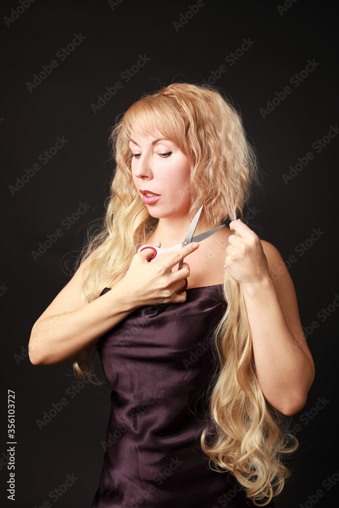 Obraz premium Blonde middle-aged caucasian woman cutting her hair with scissors and drink glass of red wine isolated on black background 