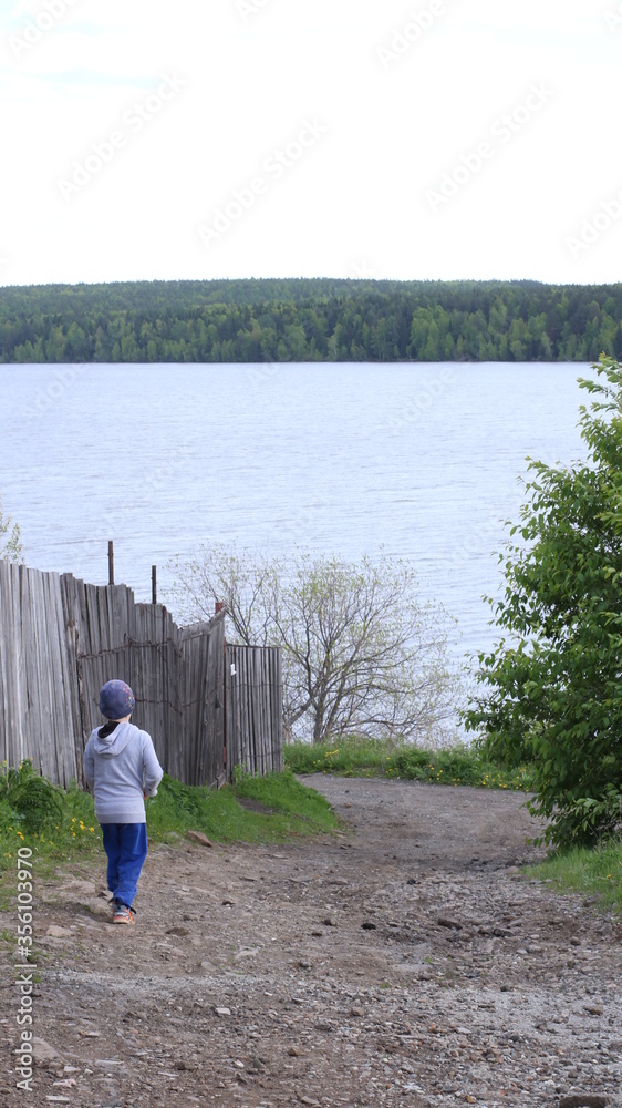 a lonely boy descending a rocky path along a wooden fence to the bank of a river or lake with a forest on the opposite side