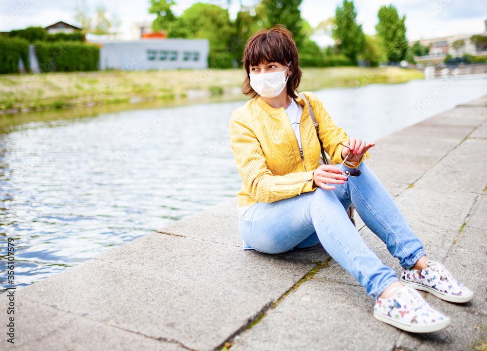 Pretty youthful middle-aged woman in white medical mask in casual clothes sits on steps of city park on sunny spring warm day. Concept is protected from bacteria and viruses in an urban environment. Pretty youthful middle-aged woman in white medical mask in casual clothes sits on steps of city park on sunny spring warm day. Concept is protected from bacteria and viruses in an urban environment.