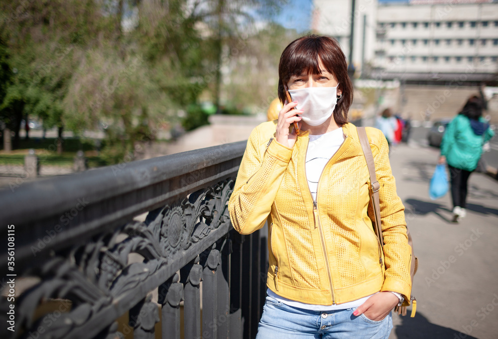 Fototapeta premium Funny middle-aged woman in a white protective mask talking on a smartphone while walking around the city on a warm spring day. Coronovirus pandemic and remote communication concept
