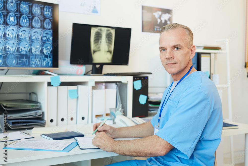 Portrait of handsome mature doctor looking at camera while posing at workplace in modern clinic interior with CT scans in background, copy space Portrait of handsome mature doctor looking at camera while posing at workplace in modern clinic interior with CT scans in background, copy space