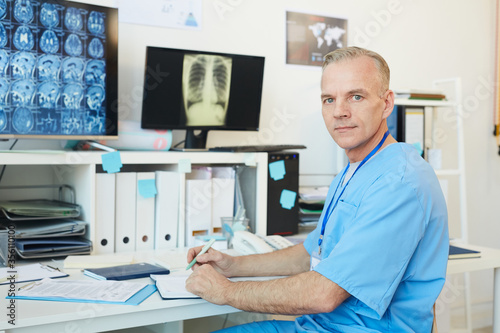Portrait of handsome mature doctor looking at camera while posing at workplace in modern clinic interior with CT scans in background  copy space