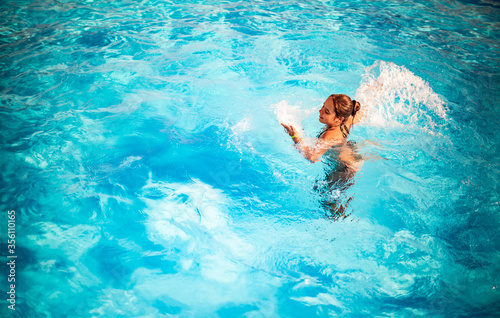 Top view pretty young girl happily splashing in blue clear water in the pool under the rays of bright sunlight. Concept of relaxation at the hotel and at sea. Advertising space