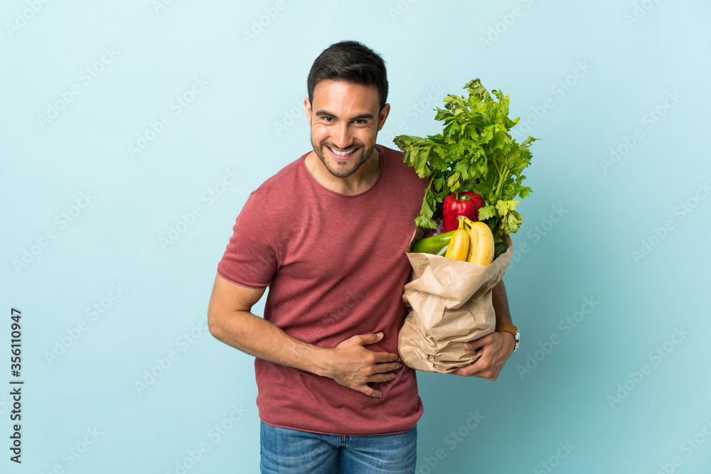 Young caucasian man buying some vegetables isolated on blue background laughing Young caucasian man buying some vegetables isolated on blue background laughing