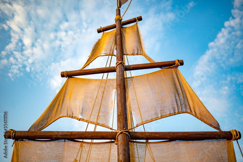 Bottom view of a ship mast with beige sails swings against a blue sky with sunny sunny summer warm day clouds. Marine and adventure concept