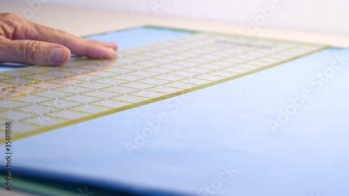 Woman tailor cutting blue cotton fabric using a rotary cutter on a cutting pad. Hand holding a ruler as a knife cuts a piece of fabric.