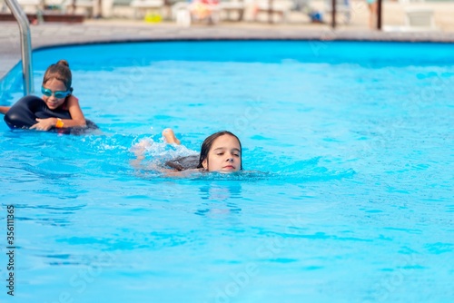 Two little sisters girls are swimming in a large pool with clear blue water of mine near the hotel on the background of the sea and the beach. Concept vacation tropical hot country with children © YouraPechkin