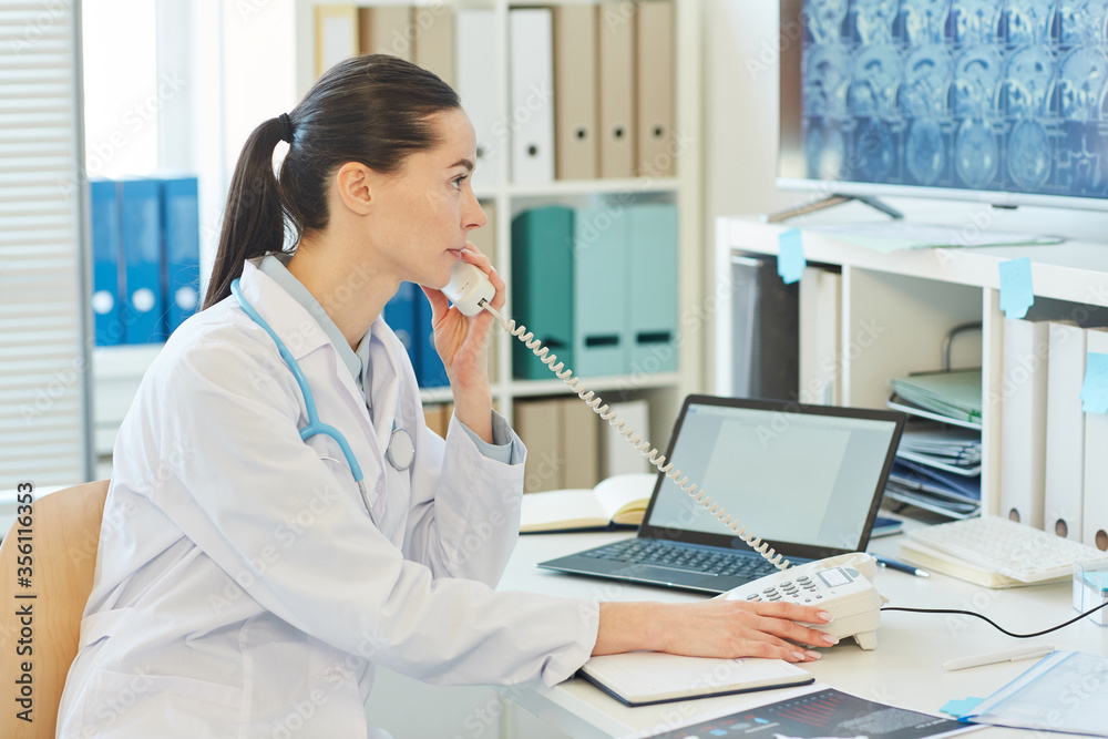 Side view portrait of young female doctor speaking by phone while sitting at desk and working in clinic or hospital, copy space Side view portrait of young female doctor speaking by phone while sitting at desk and working in clinic or hospital, copy space