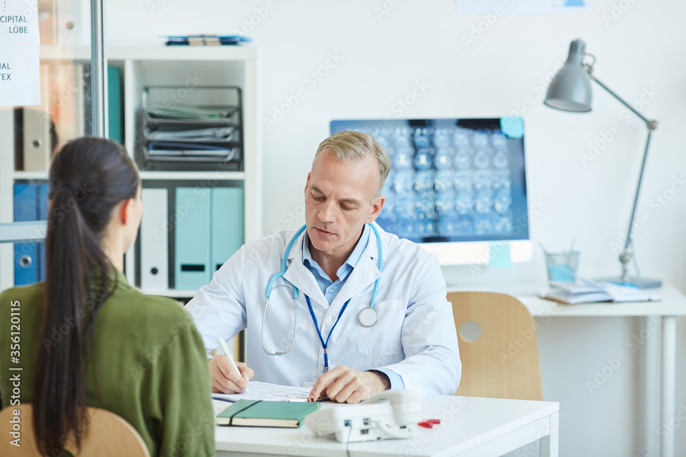 Portrait of mature male doctor writing on clipboard while sitting at desk and consulting female patient in modern clinic, copy space