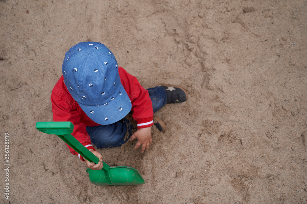 one-year-old boy with a cap, red jacket, blue pants and sneaker sits in the sand and digs with a green shovel one-year-old boy with a cap, red jacket, blue pants and sneaker sits in the sand and digs with a green shovel