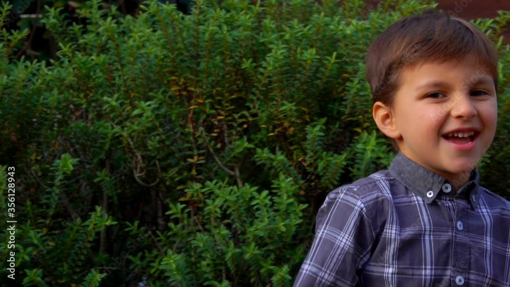 Happy boy is laughing loudly and swaying from side to side outdoors on the background of green bush Happy boy is laughing loudly and swaying from side to side outdoors on the background of green bush