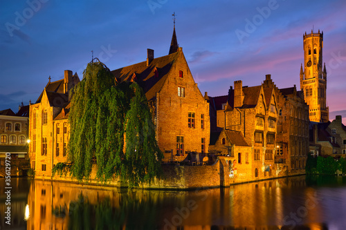 Famous view of Bruges tourist landmark attraction - Rozenhoedkaai canal with Belfry and old houses along canal with tree in the night. Brugge, Belgium