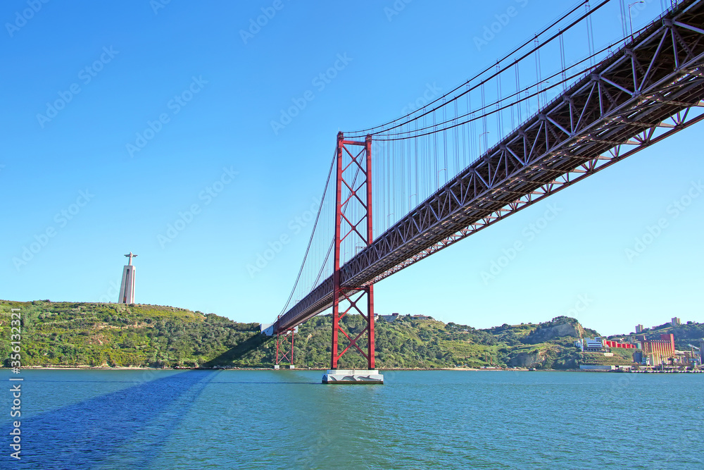 Naklejka premium 25 de Abril suspension Bridge connecting the city of Lisbon, to Almada & the statue of Christ in the background standing high above the southern banks of the Tejo Estuary, Lisbon, Portugal.