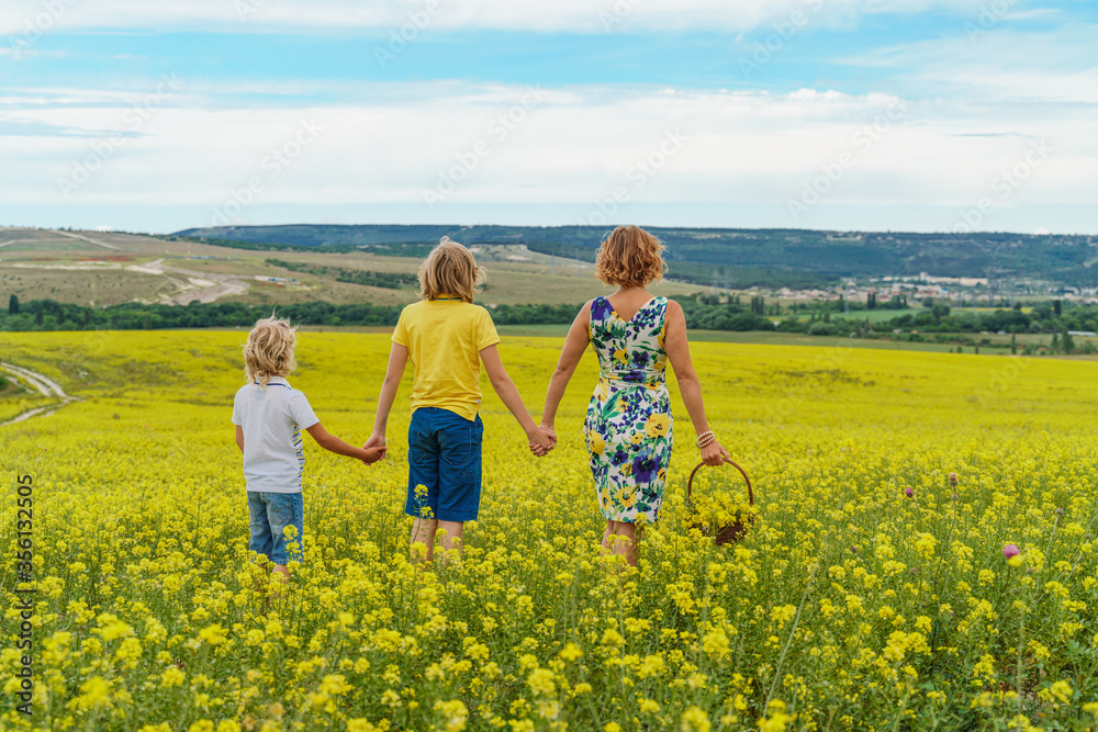 Back view of a beautiful family, a mother and two sons on a yellow flowering field of rapeseed, holding hands. Walking in the open air in nature. Mother in a dress, sons in t-shirts and shorts. Back view of a beautiful family, a mother and two sons on a yellow flowering field of rapeseed, holding hands. Walking in the open air in nature. Mother in a dress, sons in t-shirts and shorts.