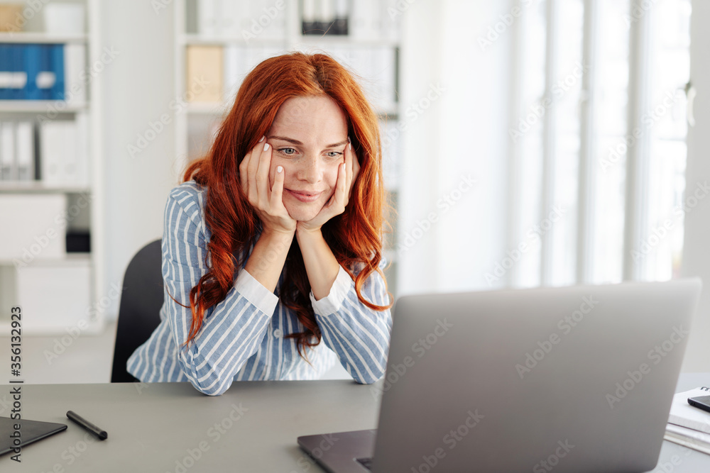 Red-haired woman in office looking at laptop Red-haired woman in office looking at laptop