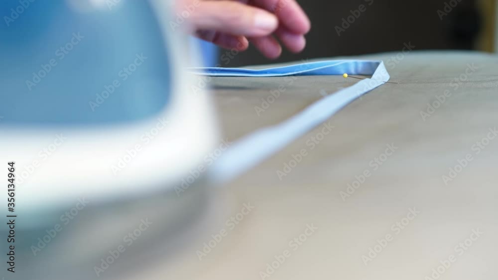 Elderly woman ironing a cotton face mask strap with a yellow pin laying on an ironing board.