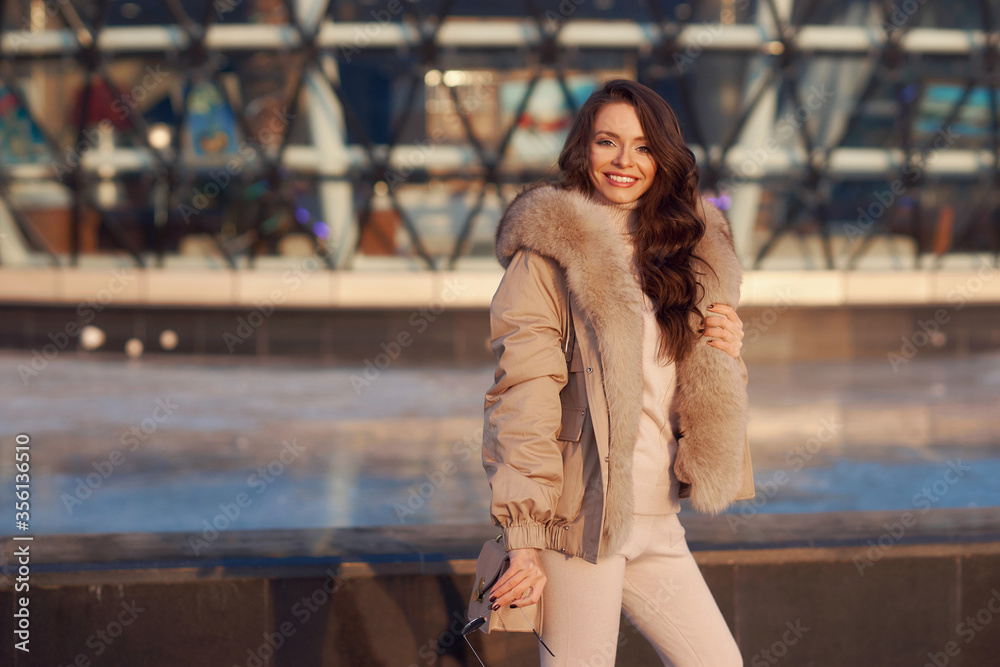 Fototapeta premium Stylish young woman wearing casual winter clothes and coat with fur hood standing or walking at city street on a sunny day. Fashionable girl