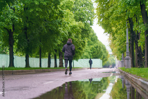 Fototapeta Naklejka Na Ścianę i Meble -  Gorky Park (Neskuchny Garden) - popular place for walking of citizens. Rainy summer day. Coronavirus pandemic. Sports allowed by the authorities. Young man in black clothes jogging