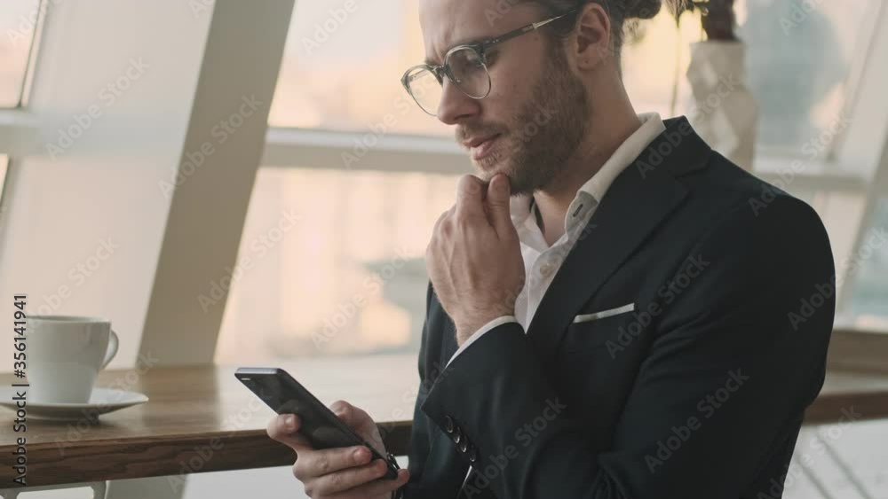 A serious thoughtful young bearded businessman is using his smartphone while drinking coffee in a city cafe indoors A serious thoughtful young bearded businessman is using his smartphone while drinking coffee in a city cafe indoors