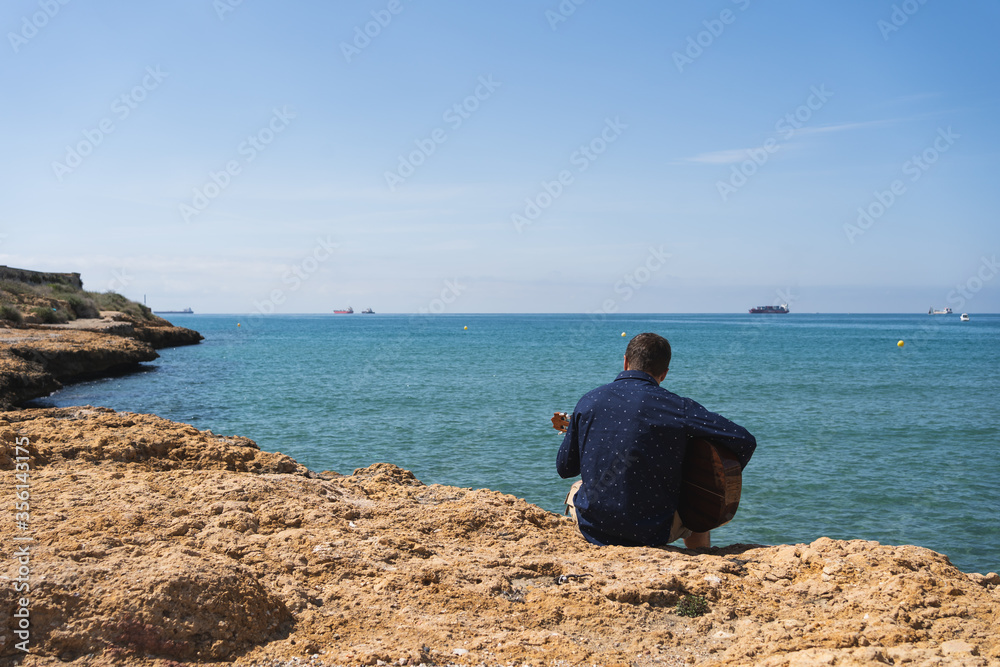 Man playing guitar sitting and facing the sea Man playing guitar sitting and facing the sea