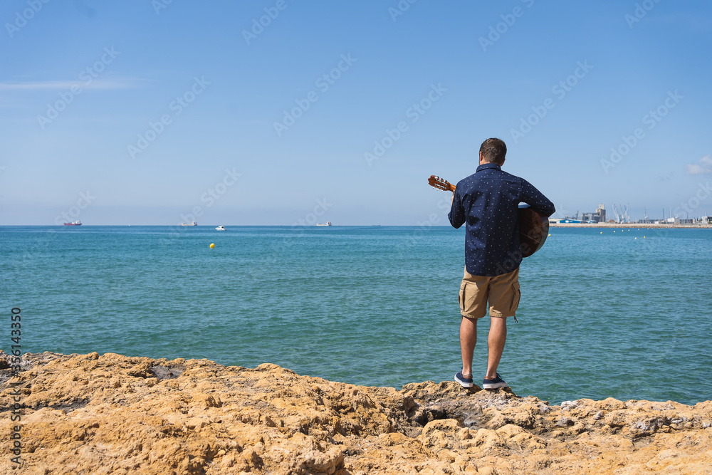 Young man with a printed shirt on his back playing a Spanish guitar standing in front of the sea