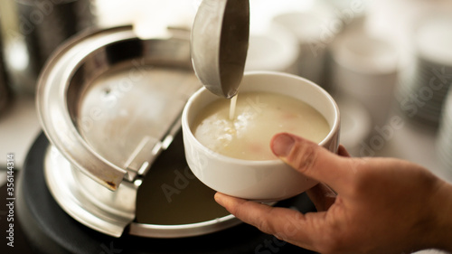 a mans hand holds a ladle and puts on the  soup ,A man pouring  soup into white dish from  tureen photo