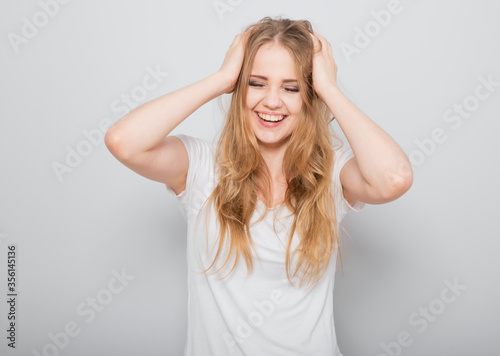 Pretty toothy laughing young woman with fair blond long hair in casual dress holding the head the hands. Studio shot of good looking beautiful woman isolated against grey studio wall